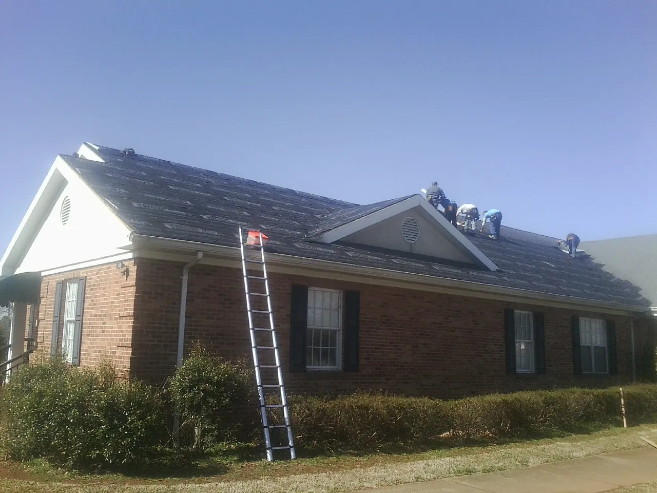 Crew tearing shingles off a brick ranch roof with a ladder set — Metal Roof Repair work by our crew in Carleton