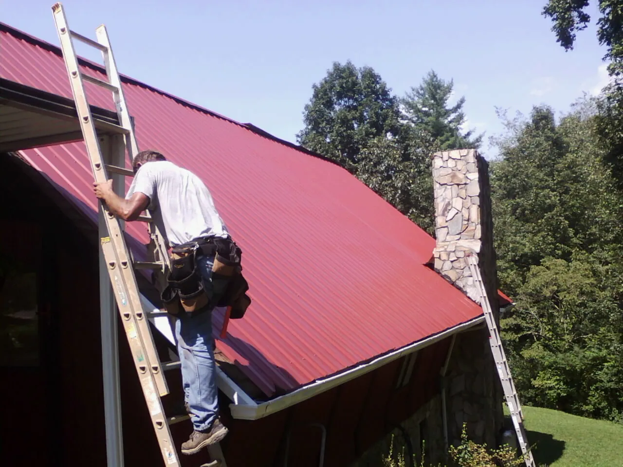 Roofer on a ladder beside a red metal roof and stone chimney — Soffit & Fascia Repair by Roofing Brothers in Carleton